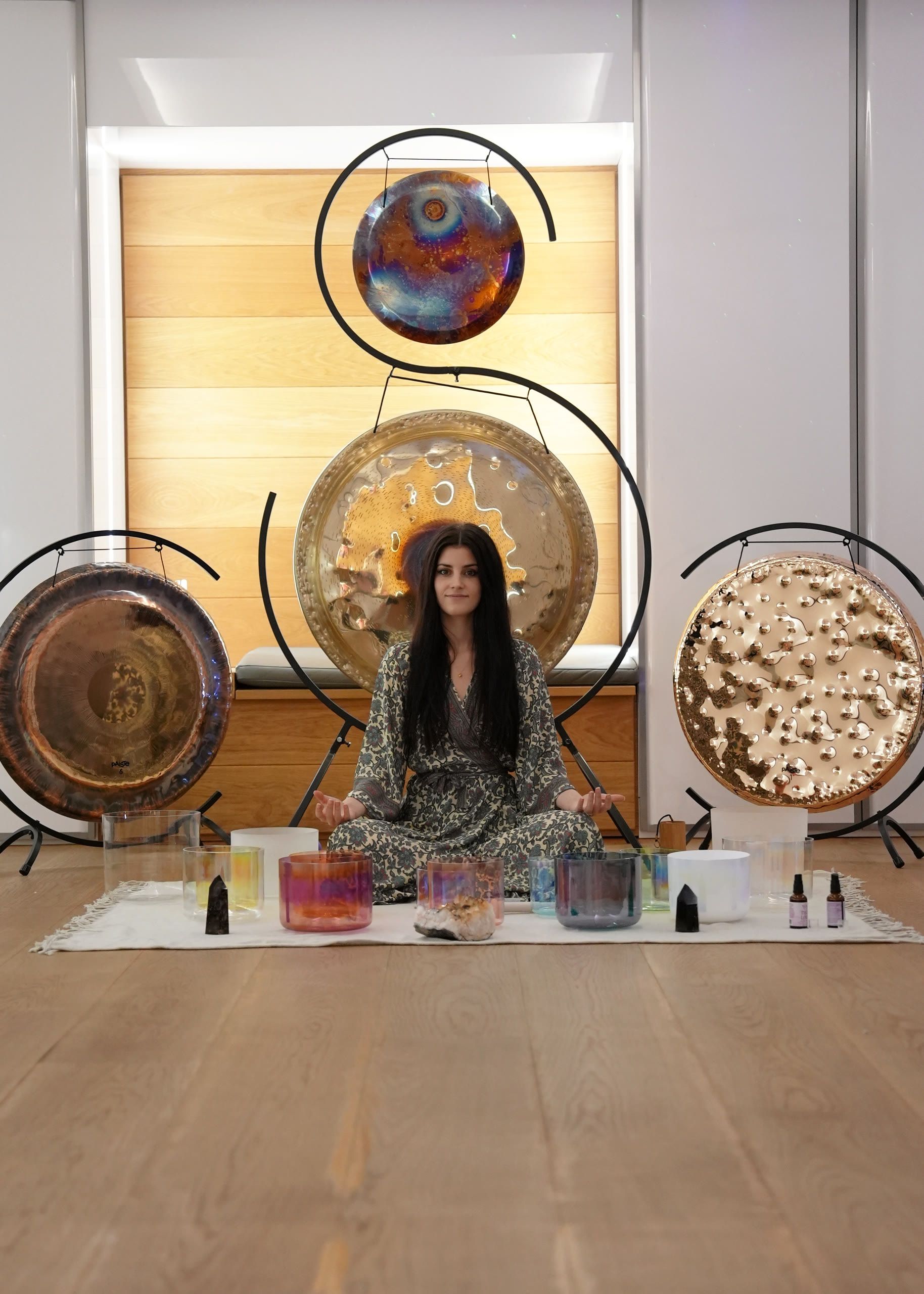 Harriet Emily sitting cross-legged among crystal singing bowls in front of three gongs in a studio
